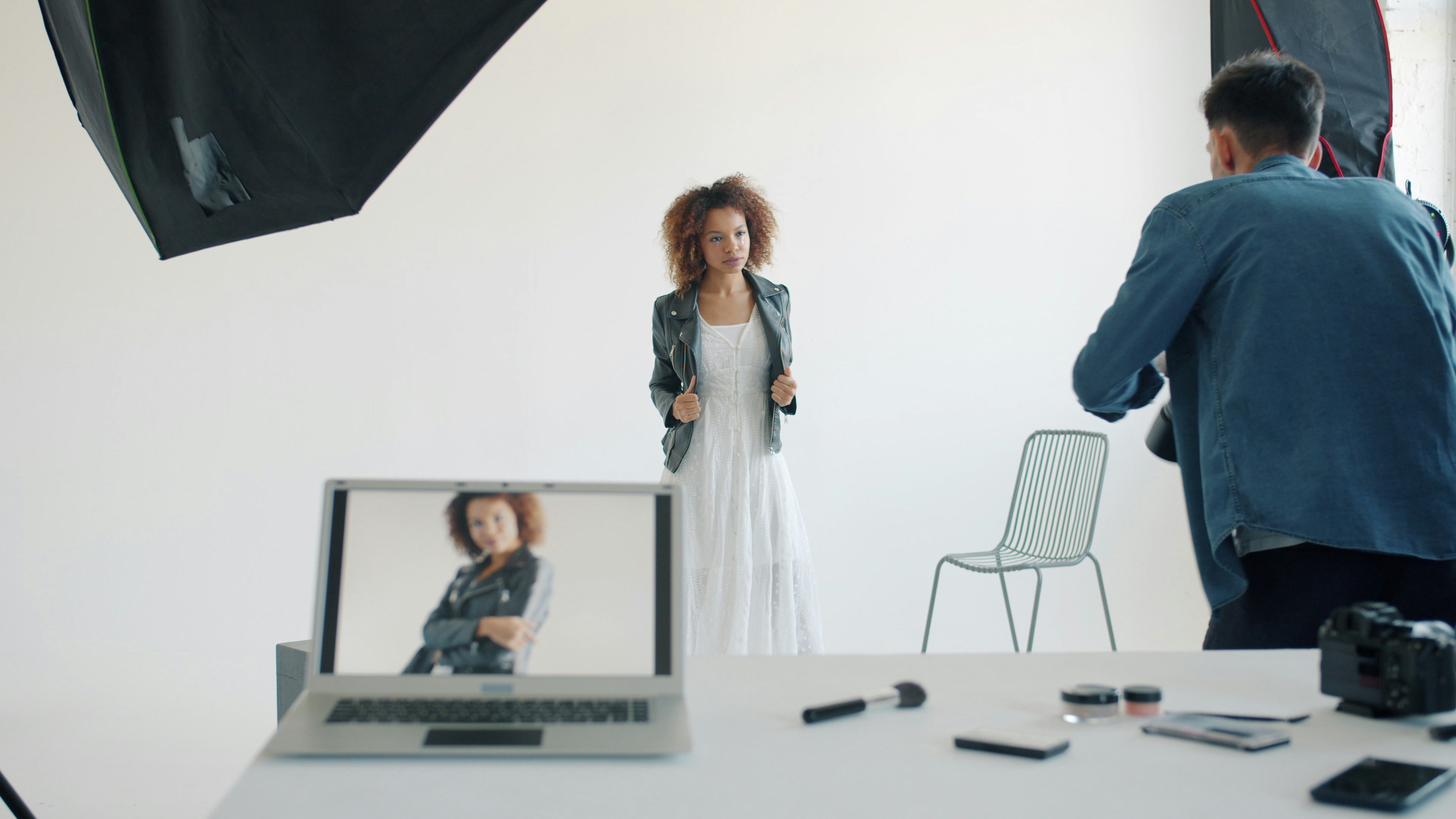 A photo studio scene showing a photographer capturing a model wearing a white dress and a black leather jacket in front of a white backdrop; a laptop on a table in the foreground displays the model’s image, with photography lights and equipment visible around the set.